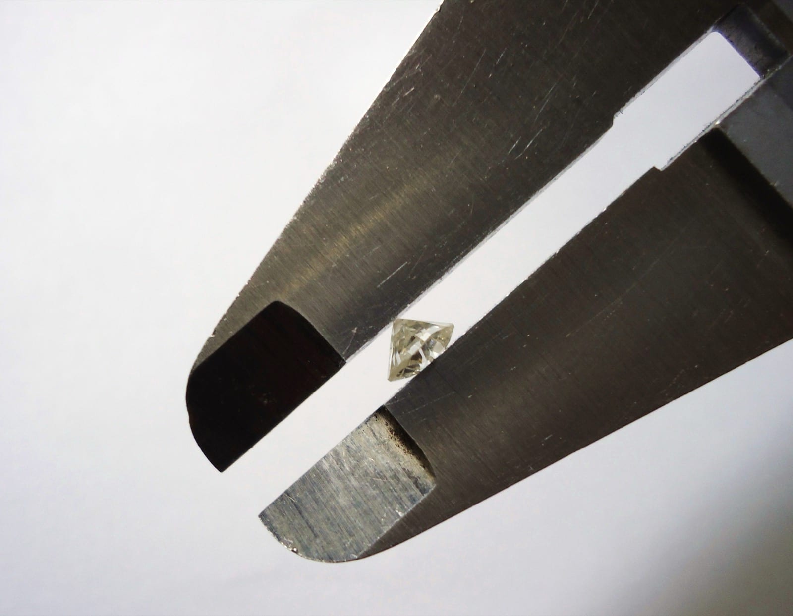 Two flat steel caliper jaws opened wide with a small faceted diamond resting between them, the stone seen from the side so its crown, girdle and pointed pavilion show against a plain pale background.