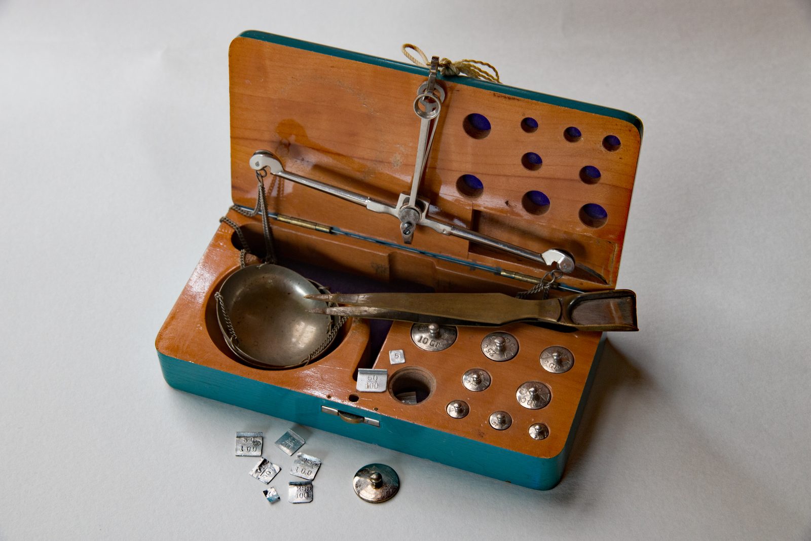 An open antique jeweler's carat balance scale with its weighing pan, a set of small brass and steel calibration weights, and tweezers laid across the case